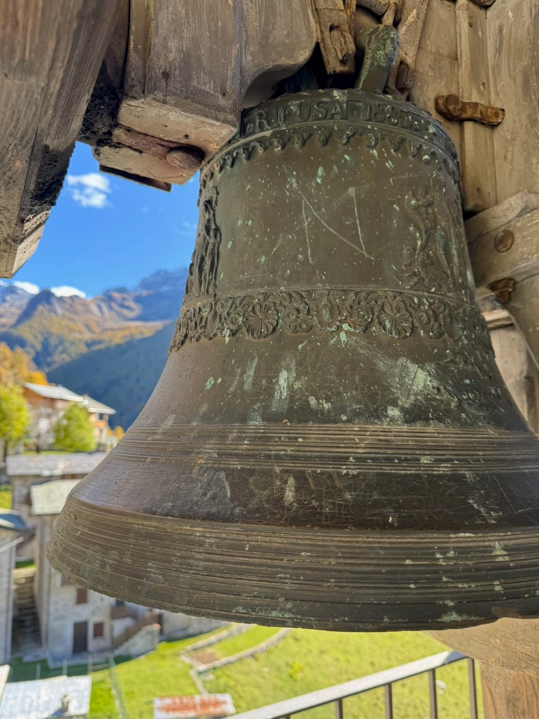 Bronzeglocke in einem Glockenstuhl, dahinter die Berge um Chiavenna